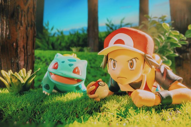 Une figurine de dresseur Pokémon allongée dans l’herbe, face à un Bulbizarre, une Poké Ball à la main